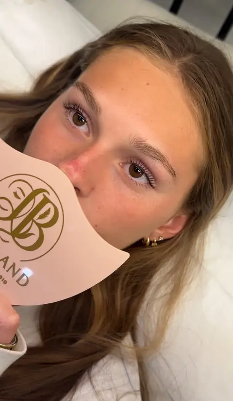 After: client with brown eyes holding a studio shield, lashes lifted and curled from the root