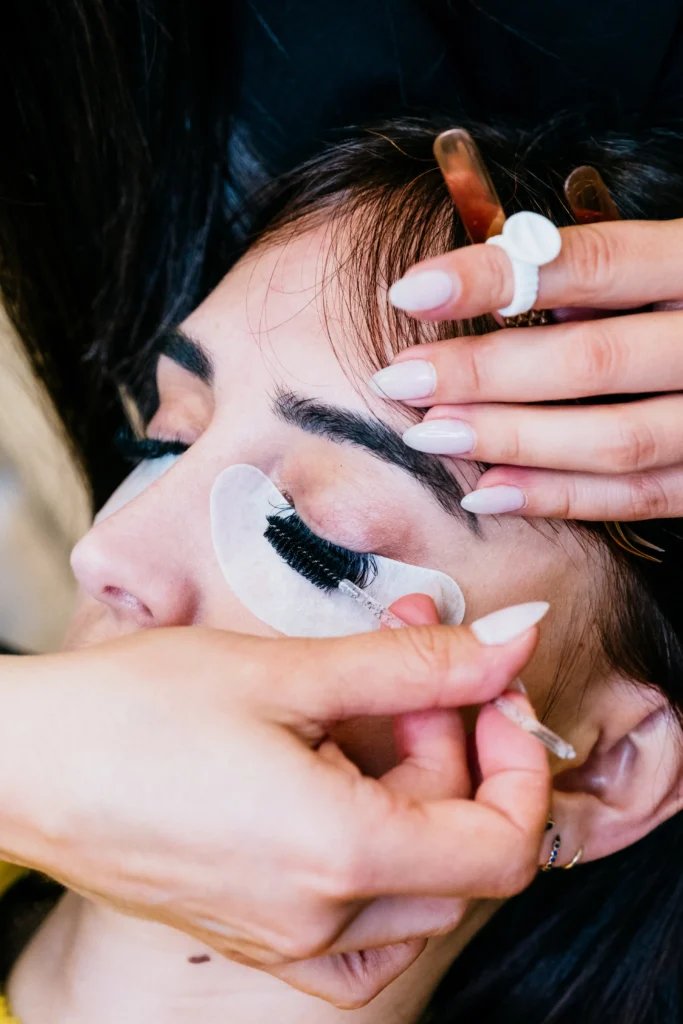 In Lakeland, a person receives eyelash extensions, with under-eye pads in place as a technician gently uses tweezers to apply individual lashes while holding their eyelid.