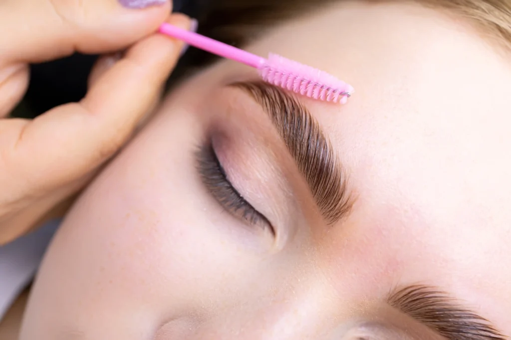 A hand uses a pink spoolie brush to groom a person's eyebrow at Lakeland; the person's eyes are closed and their eyelid is lightly made up.