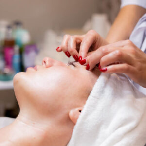 Person lying on a spa table in Lakeland with a towel wrapped around their head, receiving an eyebrow or eyelash treatment from a practitioner with red nails.