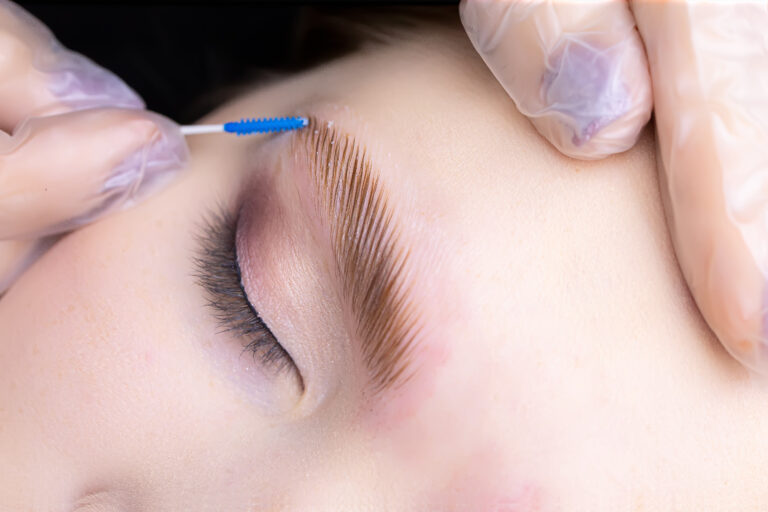 A close-up of a person’s closed eye while their eyebrow is being brushed upward with a small tool; the person, wearing protective gloves, receives a brow treatment reminiscent of Lakeland spa expertise.